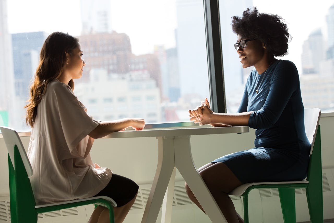 home-img Two women sitting and talking at a table with a city view from the window.